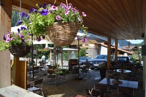 Une terrasse extérieure avec des tables et des chaises en bois, des jardinières suspendues et une vue sur les voitures et les bâtiments en arrière-plan.
