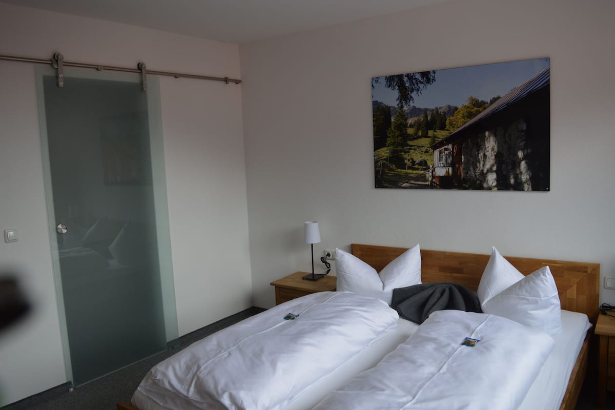 Ein Hotelzimmer mit Bett, Nachttisch, Lampe, einem Gemälde einer Berglandschaft und einer Schiebetür.