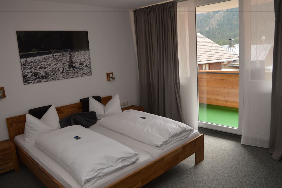 Ein Doppelbett mit weißer Bettwäsche, ein Holz-Kopfteil und ein Nachttisch mit Lampe. Ein Gemälde einer felsigen Landschaft hängt an der Wand.