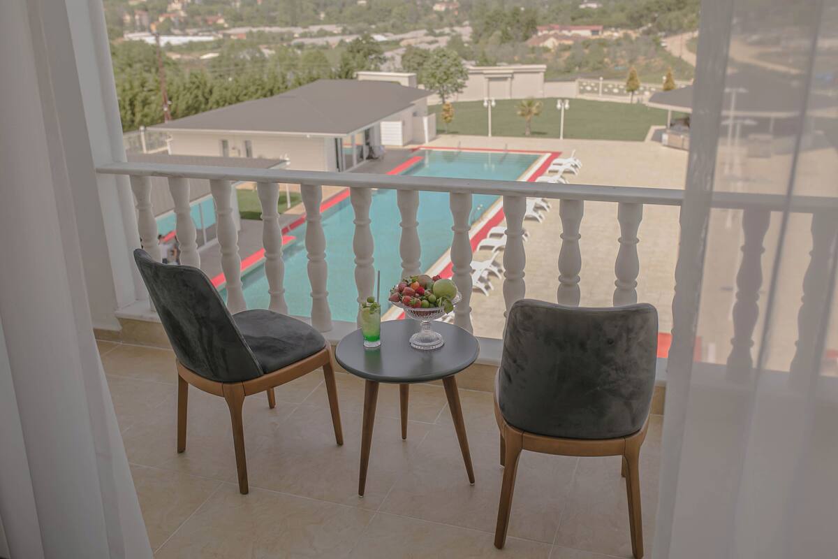 Un balcon avec deux chaises et une petite table sur laquelle se trouvent un panier de fruits et une boisson. On aperçoit une piscine à l’arrière-plan.