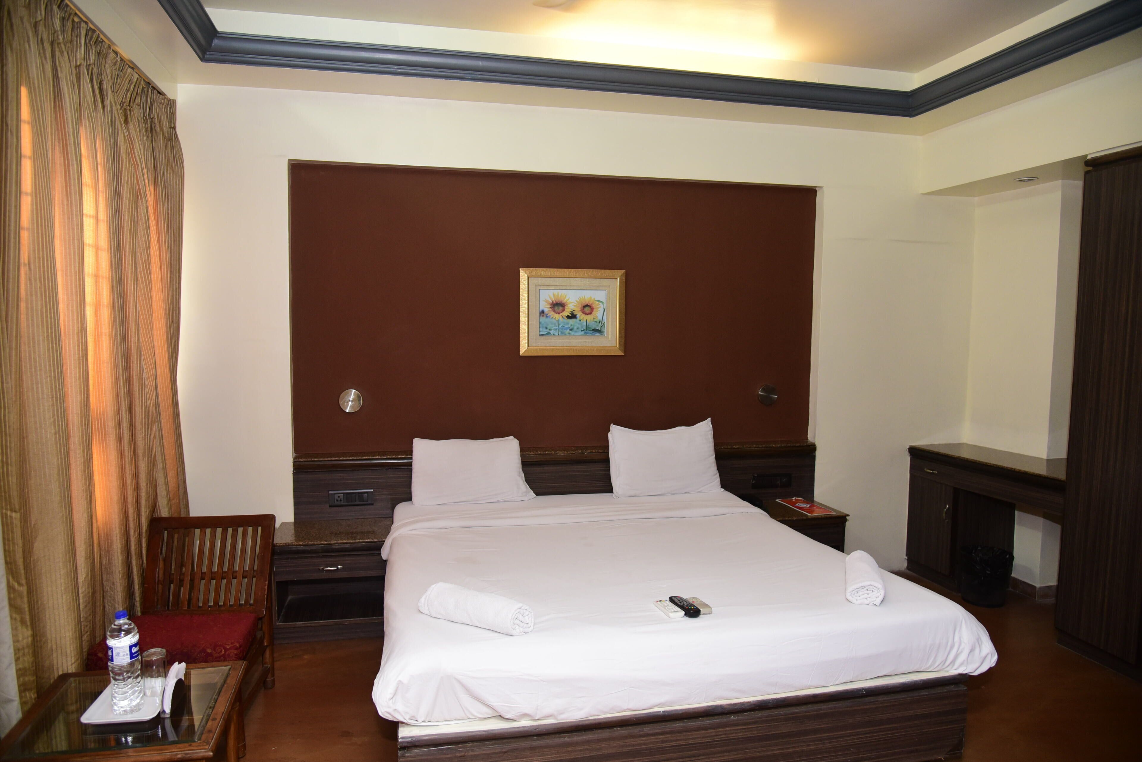 Une chambre d’hôtel avec un grand lit, une tête de lit en bois, une table de chevet, un bureau avec une chaise, un miroir et un tableau au mur.