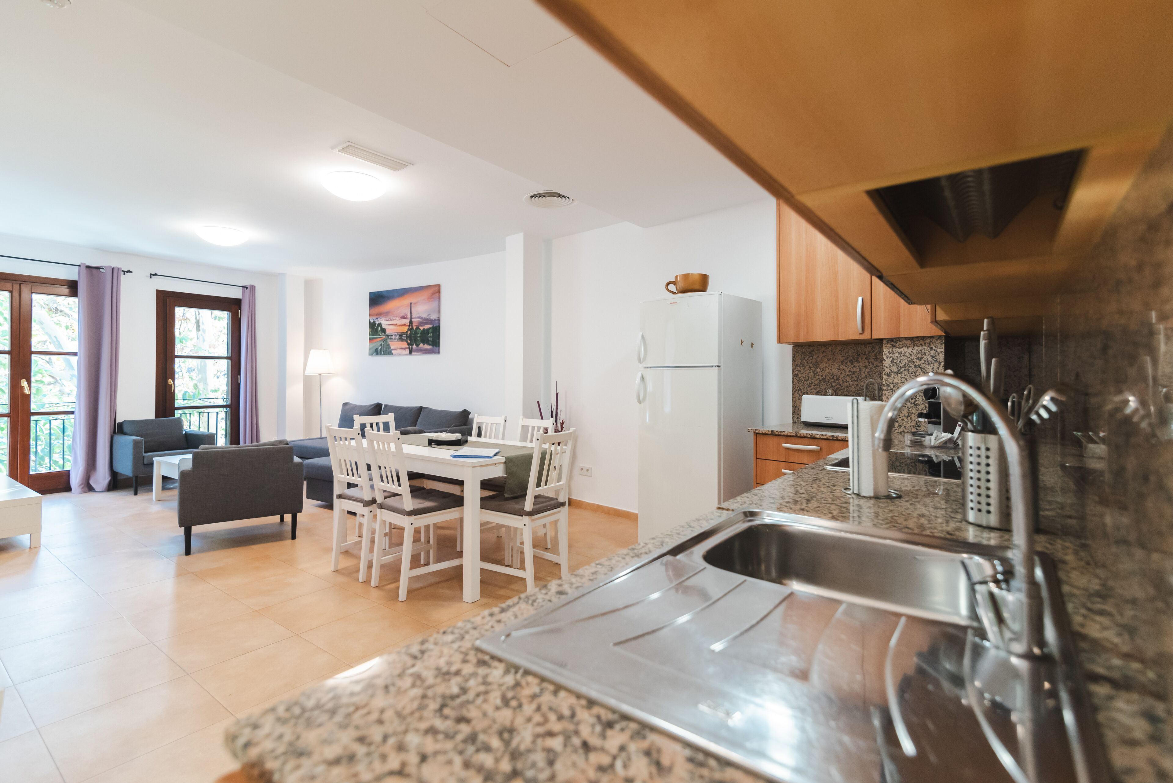 Une cuisine moderne dotée de plans de travail en granit, d’un évier en acier inoxydable et de meubles en bois. On y trouve un coin repas avec une table et des chaises, ainsi qu’un salon avec un canapé et un tableau accroché au mur.