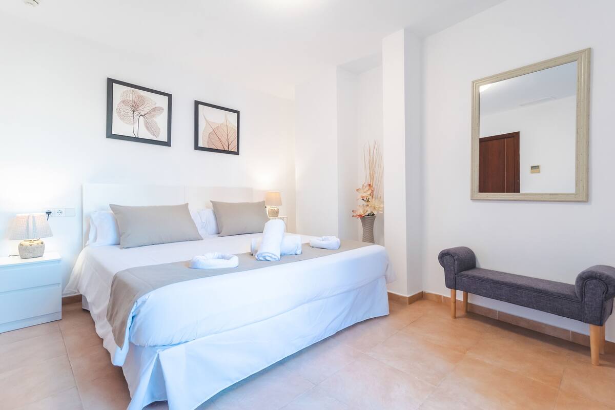 Un dormitorio ordenado con cama, mesitas de noche, un banco y cuadros en la pared.