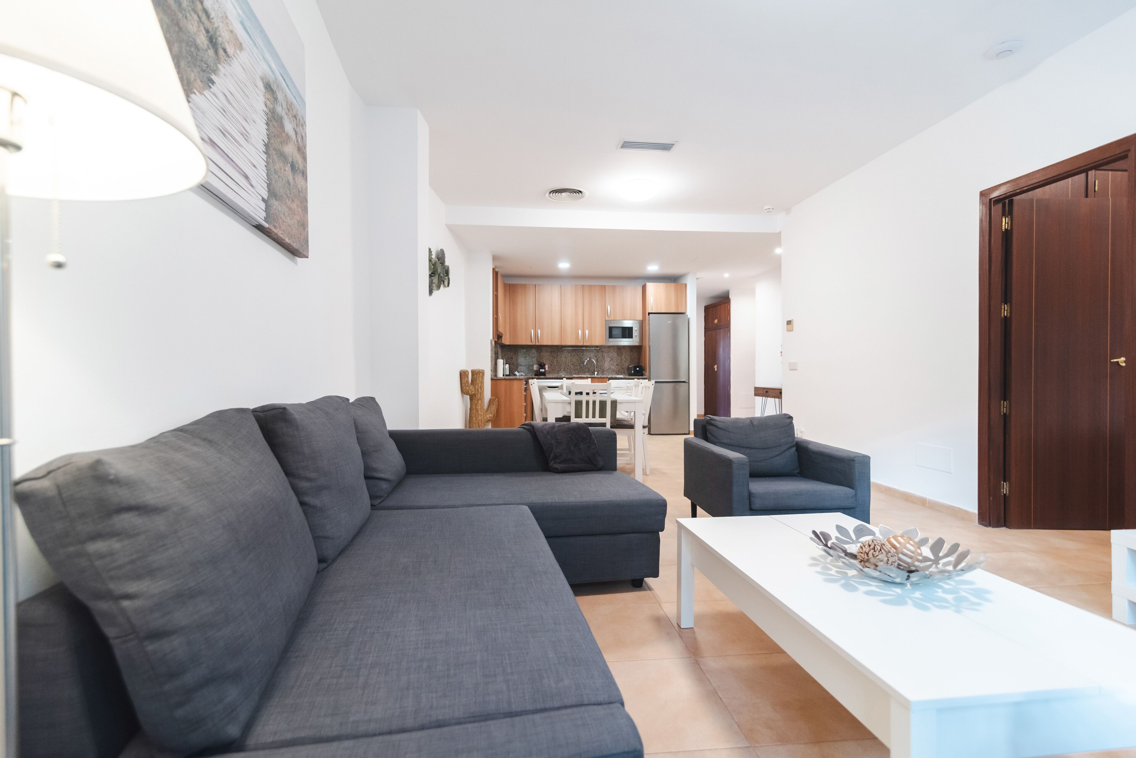 A modern living room with a grey sectional sofa, a white coffee table, and a grey armchair. A kitchen with wooden cabinets and a refrigerator is visible in the background.