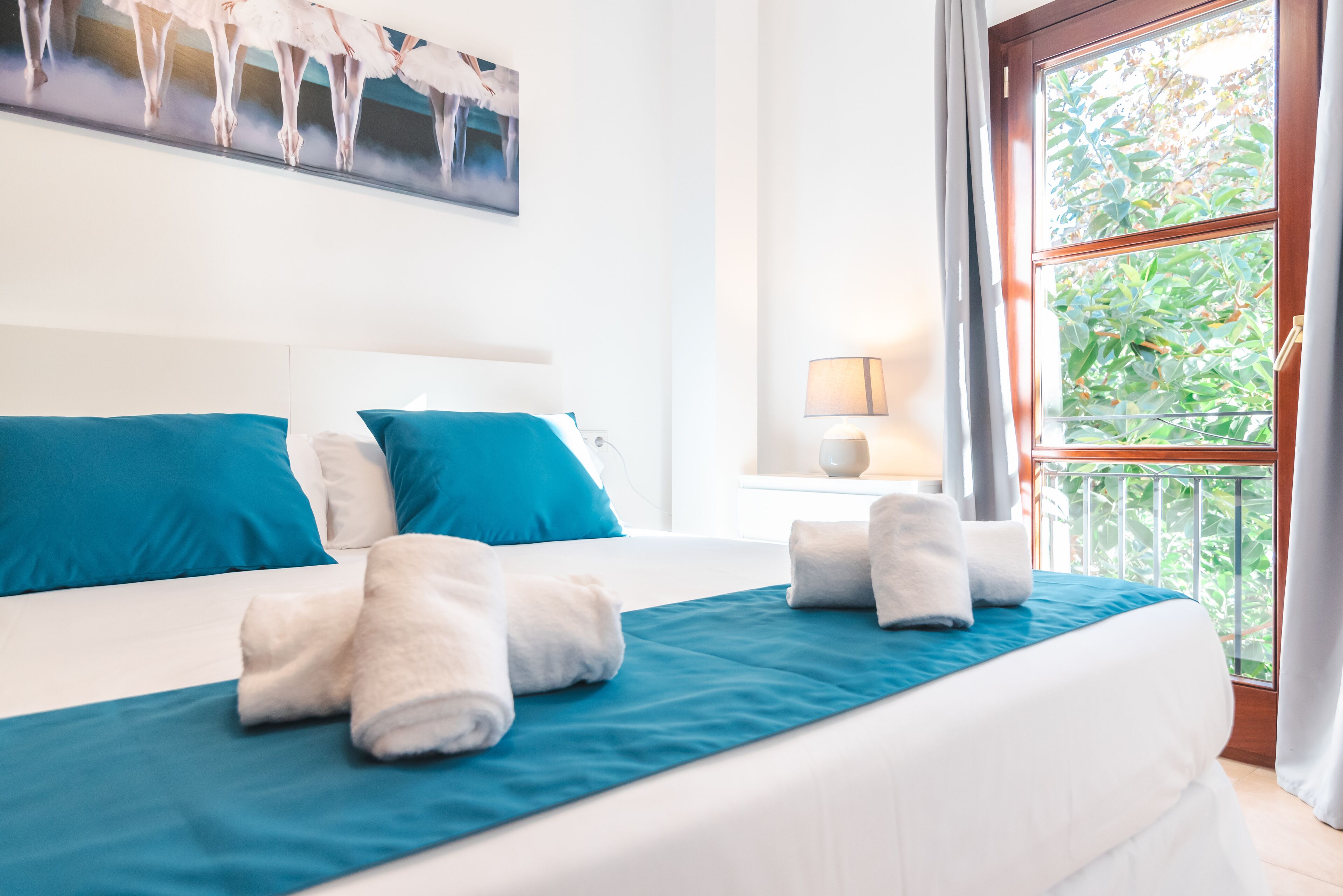 A hotel room with a large bed, blue bedding, white pillows, and a view of greenery through the window.