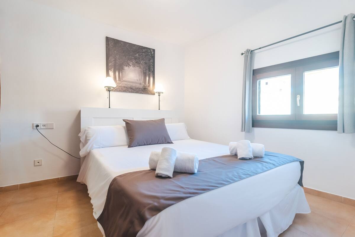 Una cama bien tendida con ropa de cama blanca y marrón, una obra de arte colgada en la pared y una ventana con cortinas.