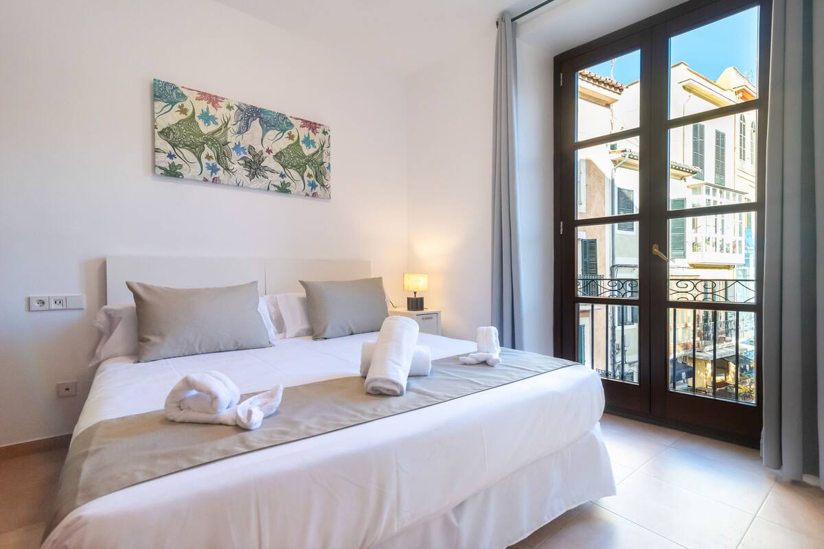 Habitación de hotel con una cama grande, una mesita de noche con una lámpara, un cuadro en la pared y vistas de edificios a través de un ventanal amplio.