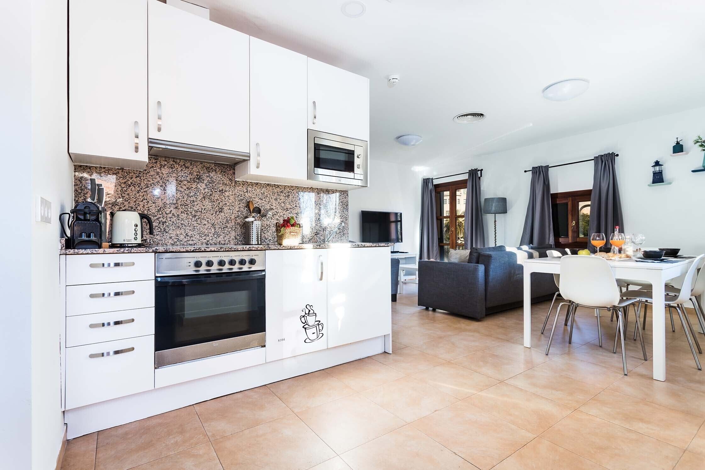 A modern kitchen with white cabinets, a built-in oven, and a microwave. There is a dining area with a white table and chairs.