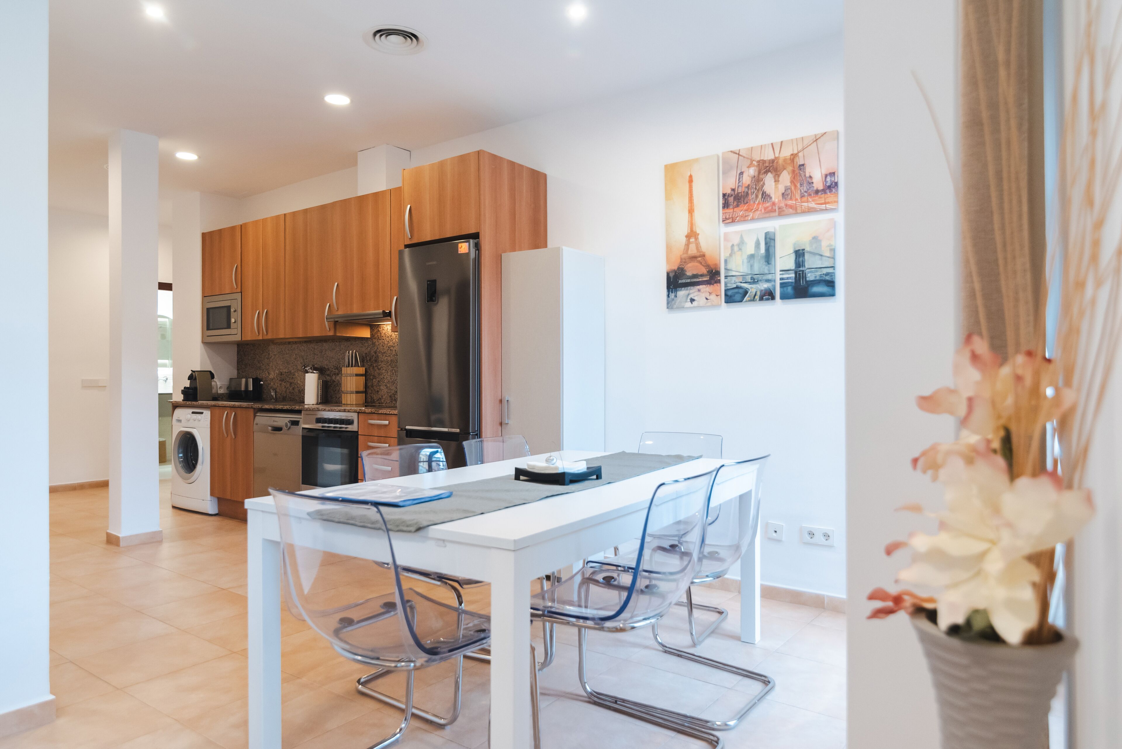 Una cucina moderna con mobili in legno, elettrodomestici in acciaio inossidabile e un'area pranzo con tavolo bianco e sedie trasparenti.