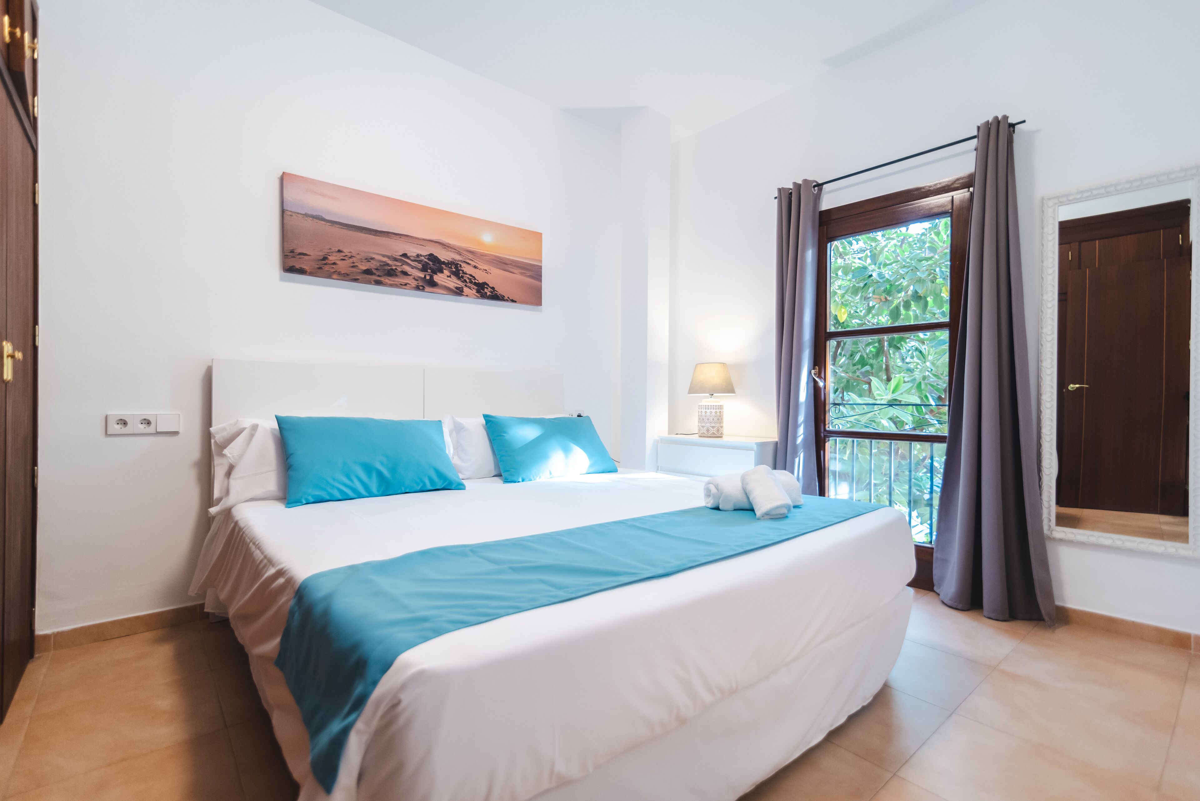 Un dormitorio con una cama grande, una mesita de noche, una lámpara, una ventana con cortinas y un cuadro en la pared.