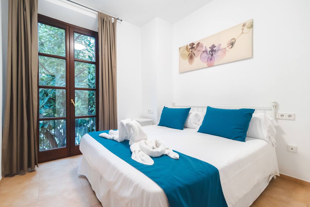 Una habitación de hotel con una cama grande, ropa de cama azul y blanca, un ventanal con cortinas y una obra de arte floral en la pared.