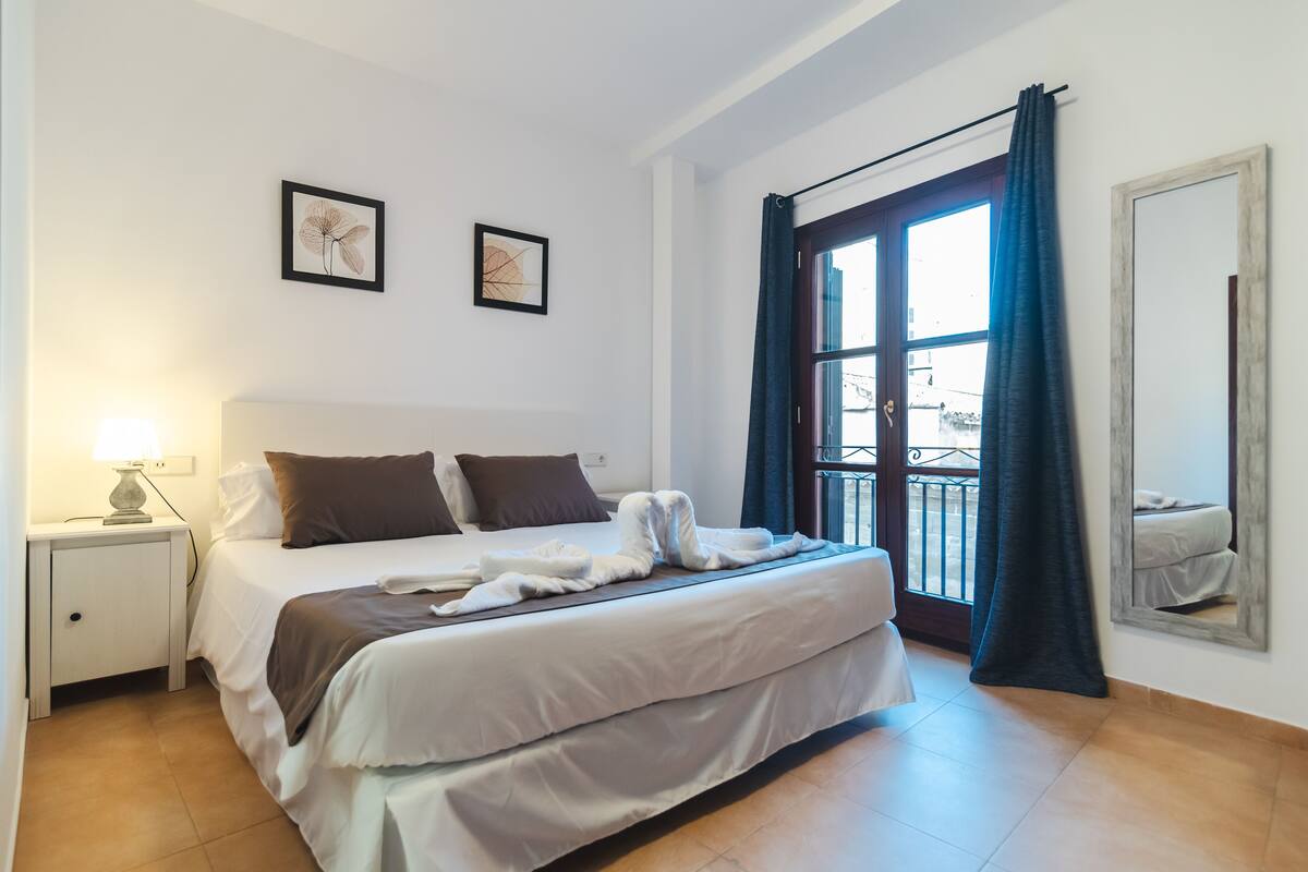 Una cama bien hecha con ropa de cama blanca y marrón, una lámpara de noche y dos cuadros en la pared.