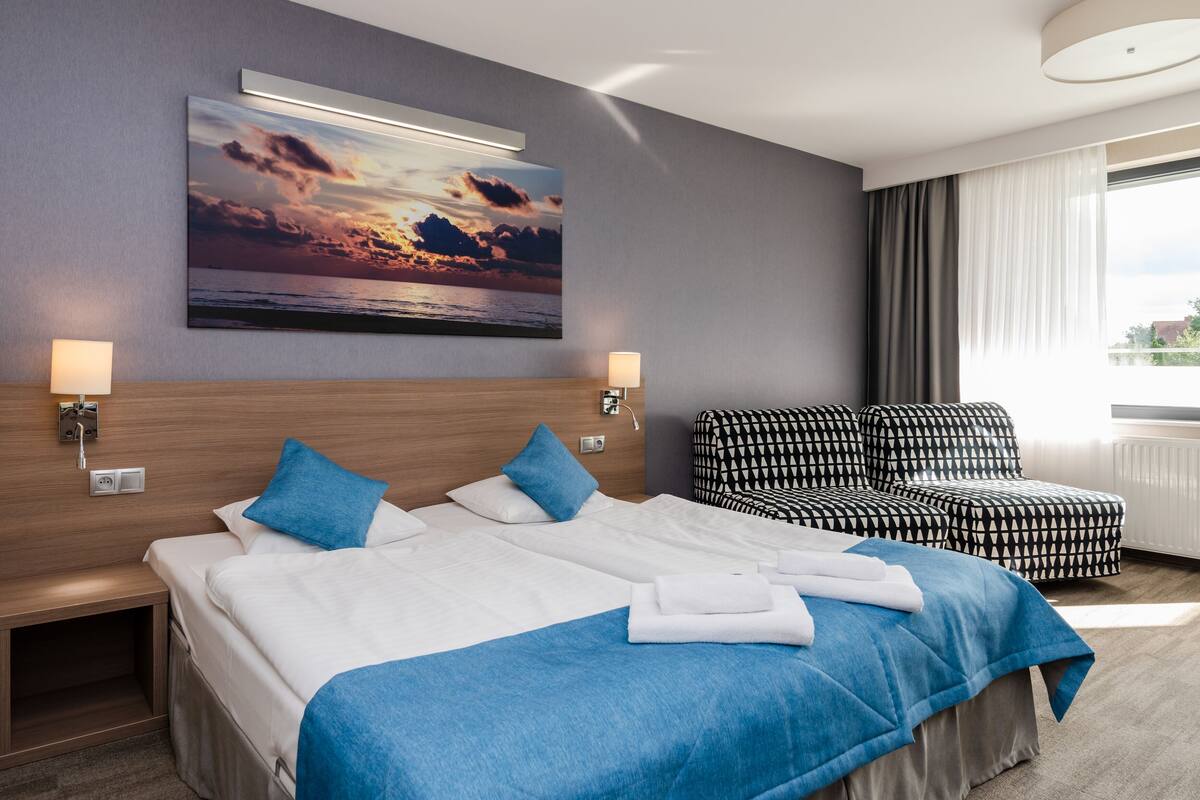 A hotel room with two beds, a large painting of a sunset, a sofa, and a view of the city.