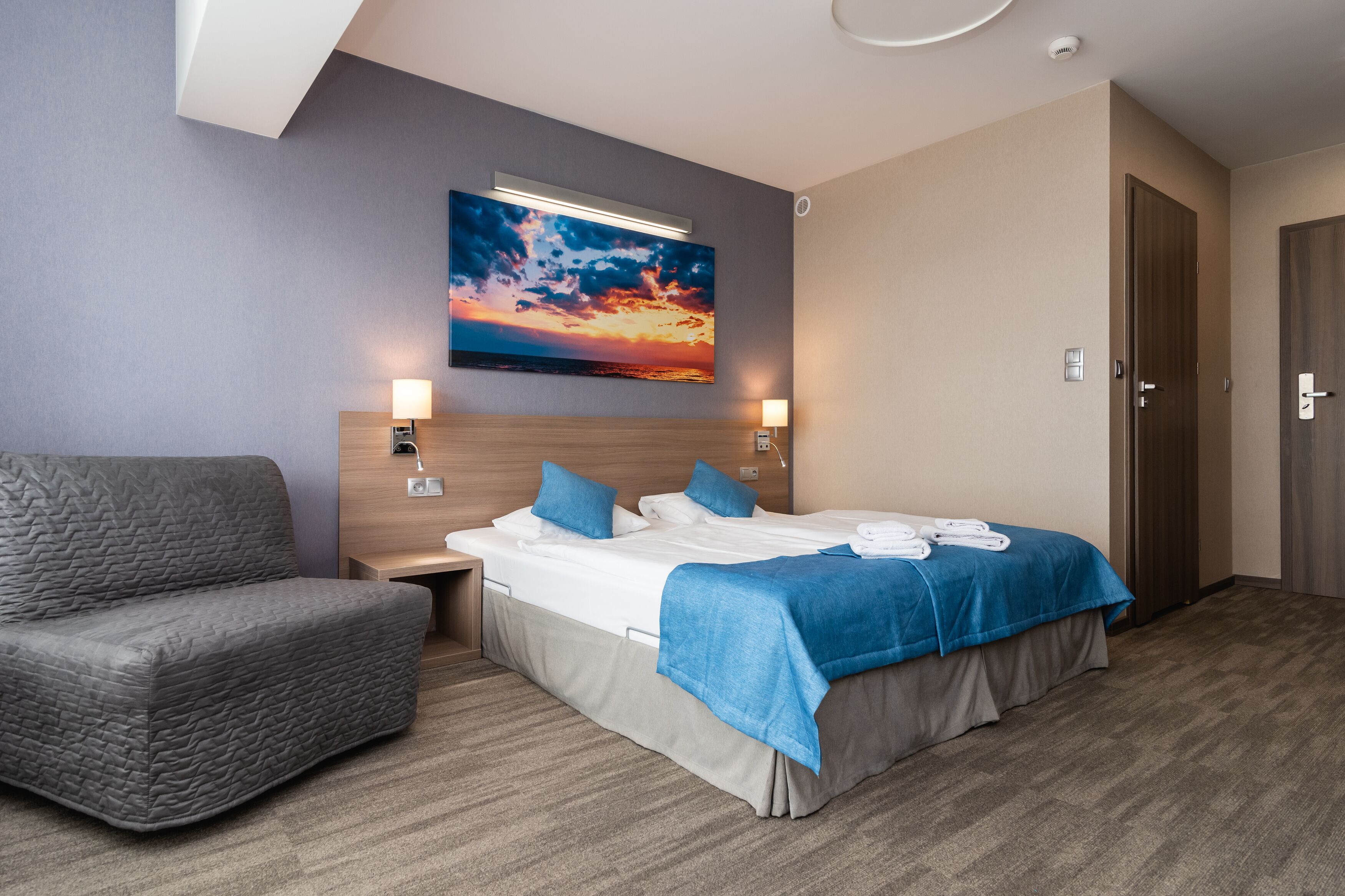 A hotel room with a bed, a sofa, a painting of a sunset, and a door.