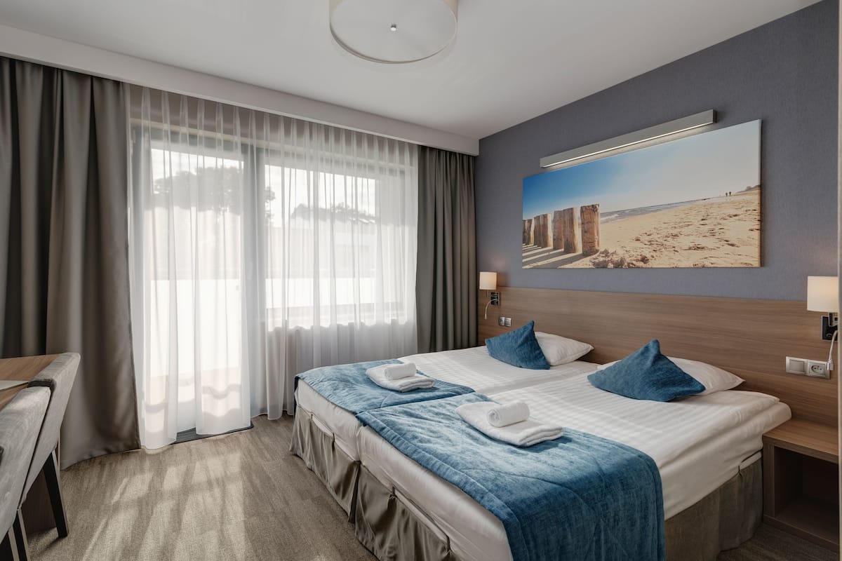 A hotel room with a bed, a desk, a chair, a large window with curtains, a wall-mounted lamp, and a painting.