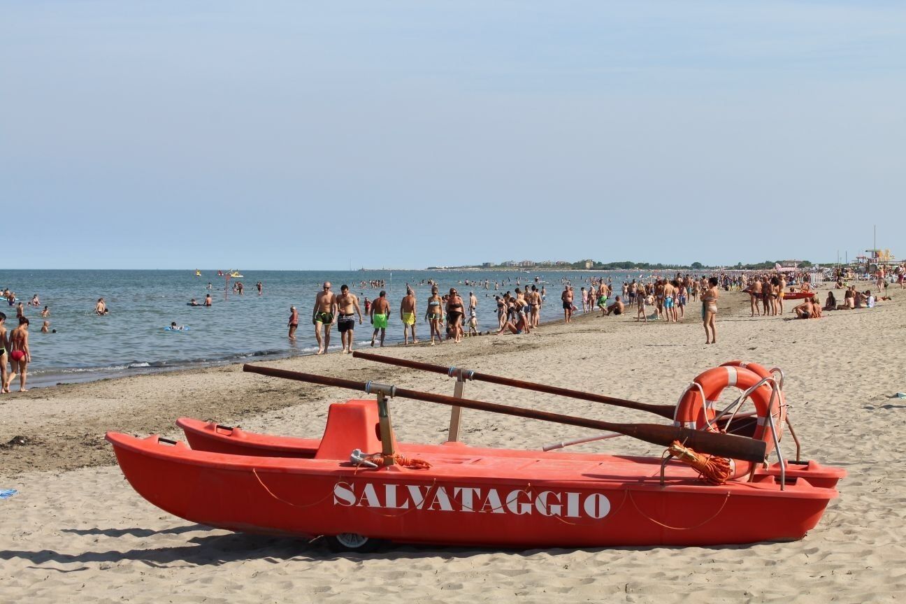 A red lifeboat labeled "SALVATAGGIO" on a beach with people in the water and on the sand.