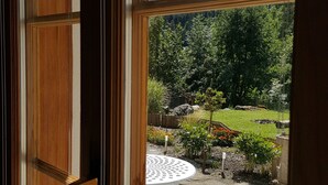 Udsigt gennem et vindue med træramme, en sten terrasse og en have med forskellige planter samt et lille bord.