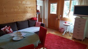 Hyggelig hytteindretning med trævæg, sofa, spisebord med tallerkener, rød stol, lille hvidt bord og fjernsyn på en kommode.