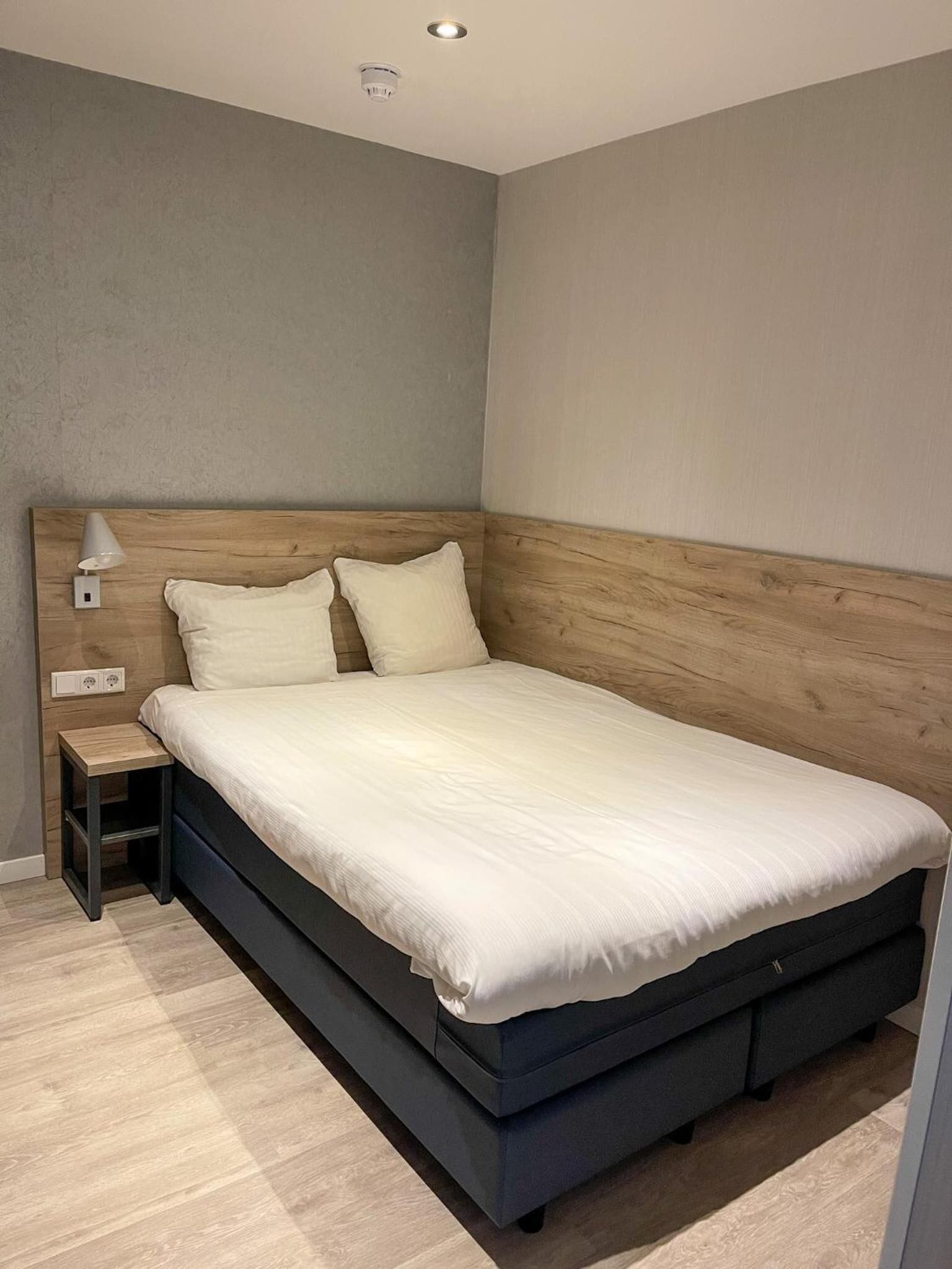 A modern bedroom with a wooden headboard, a single bed, a bedside table with a lamp, and a power outlet.