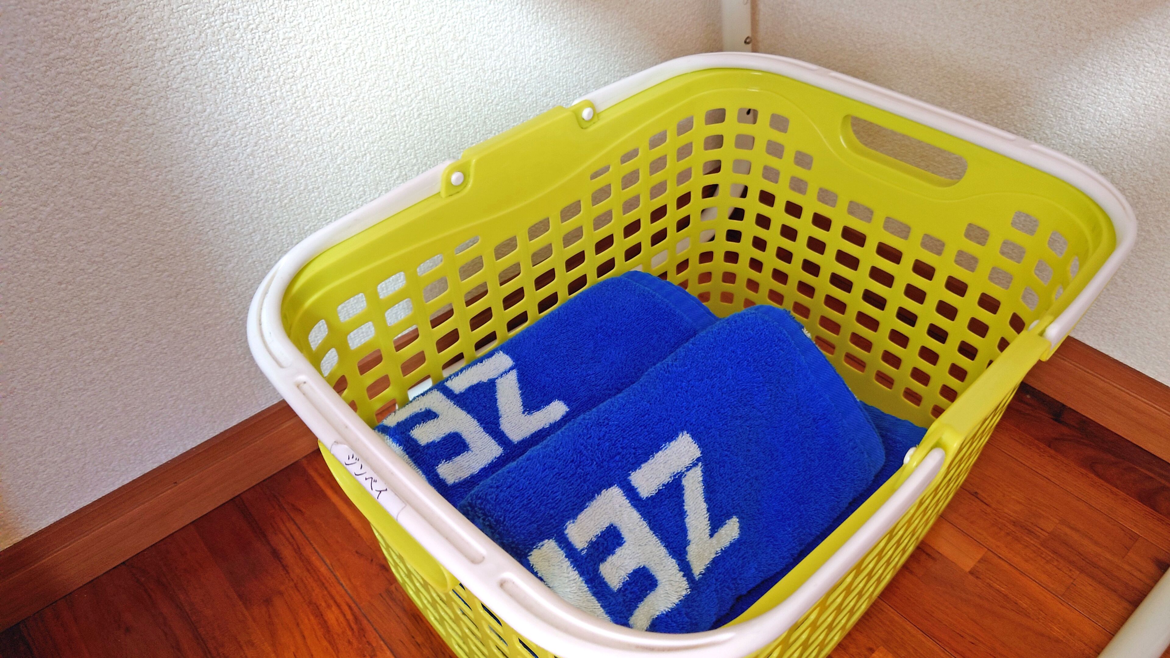 A yellow laundry basket with two blue towels inside, placed on a wooden floor against a white wall.