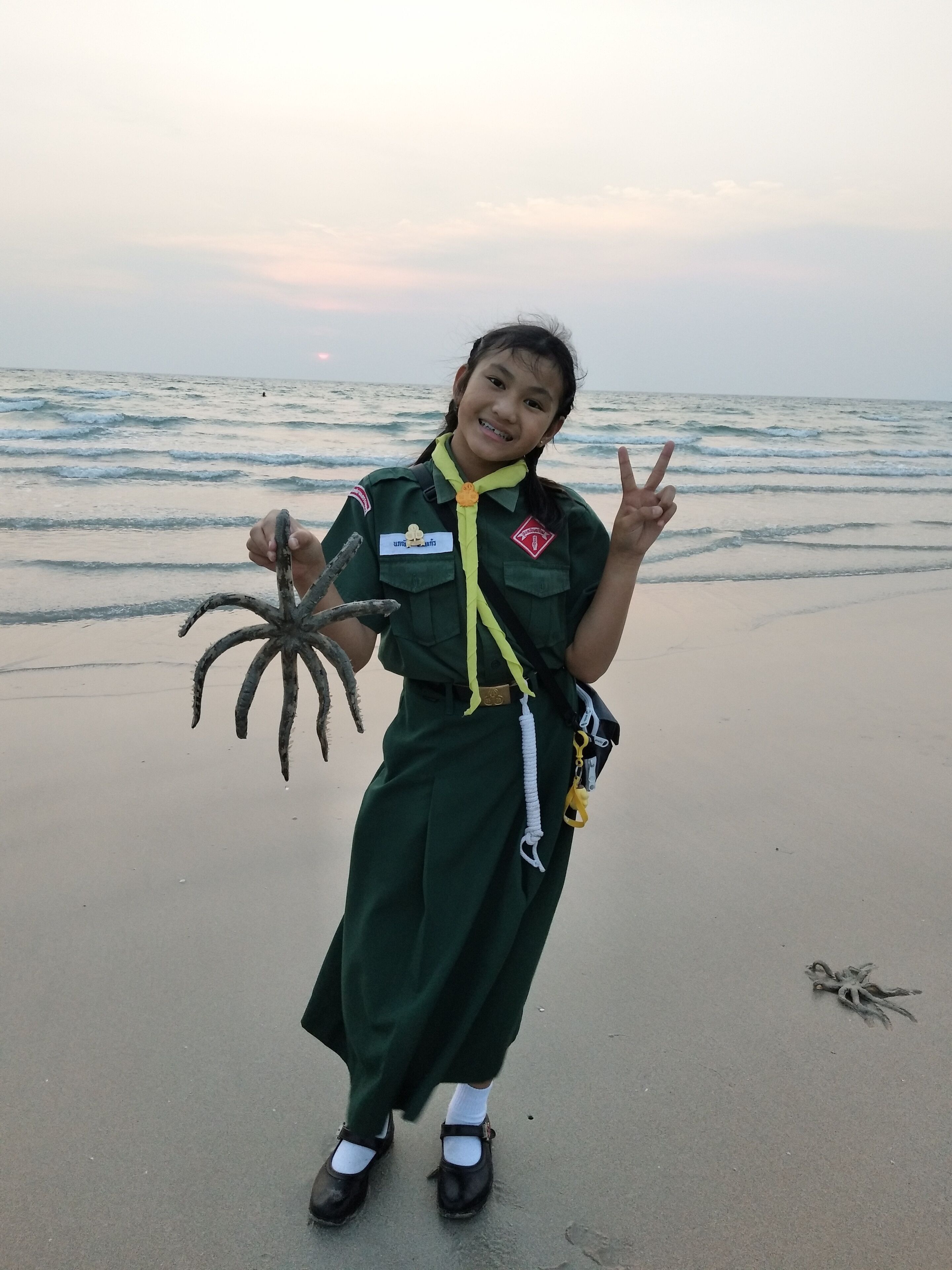 A person in a green uniform holding a starfish on a beach with the ocean in the background.