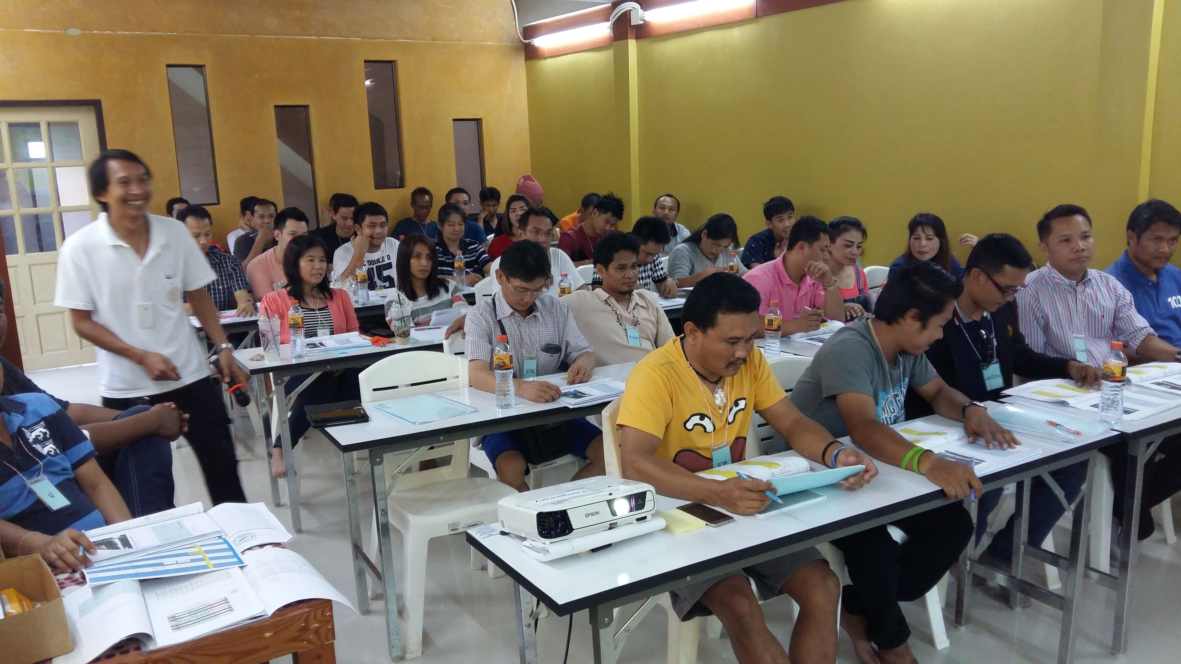 Un aula con estudiantes sentados en pupitres, algunos usando ordenadores portátiles y con papeles, y un proyector sobre uno de los escritorios.