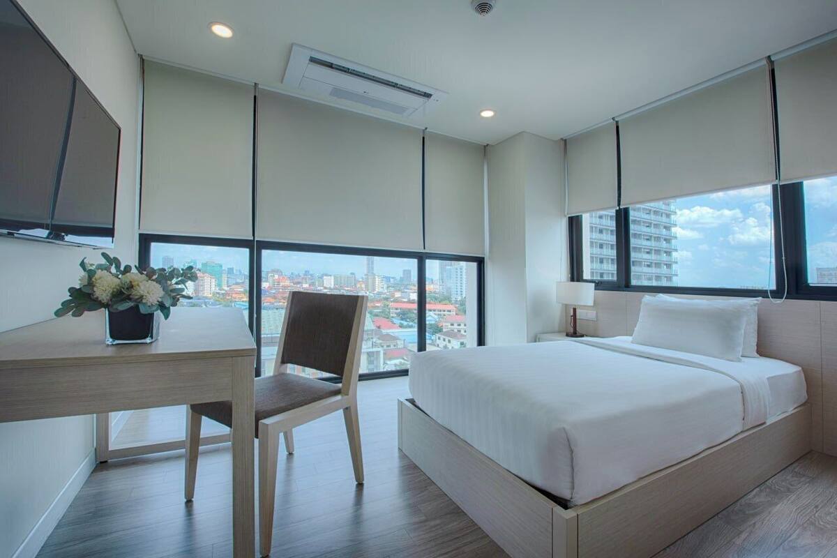 A modern hotel room with a large bed, a desk with a chair, and a view of a cityscape through large windows.