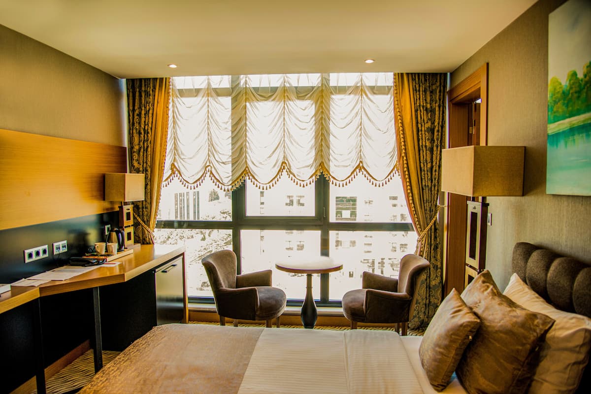 A hotel room with a bed, two armchairs, a small table, a desk, a lamp, and a window with curtains.