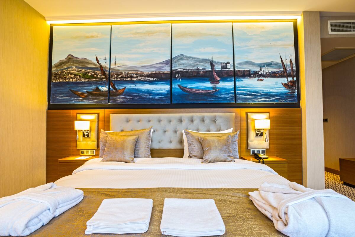 A hotel room with a large bed, two bedside tables with lamps, a headboard with a painting, and neatly folded towels on the floor.
