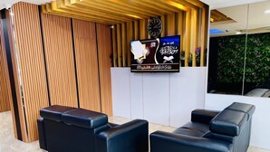 A reception area with a wooden panel wall, a mounted clock, a television displaying Arabic text, and two black leather sofas. - Cozy Hotel at KL Sentral (Kuala Lumpur)