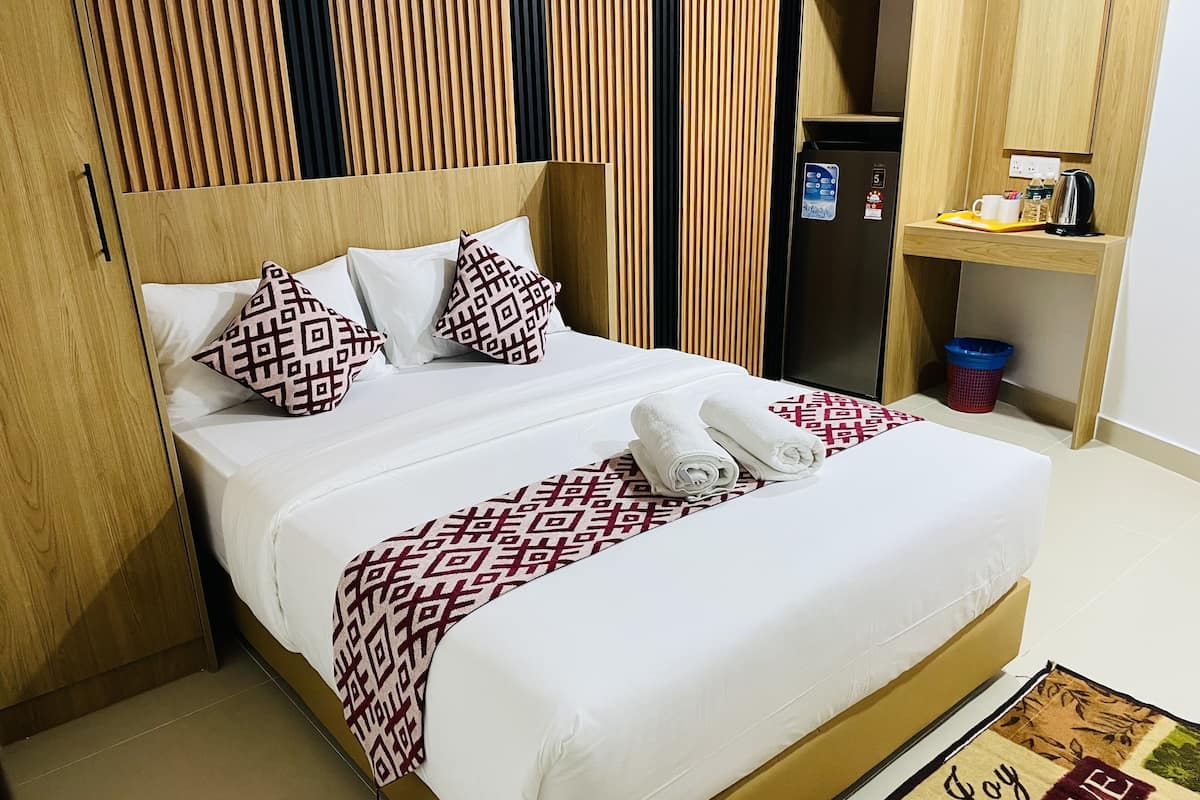 A neatly made bed with patterned pillows and a decorative throw, a small refrigerator, and a coffee maker on a wooden shelf.