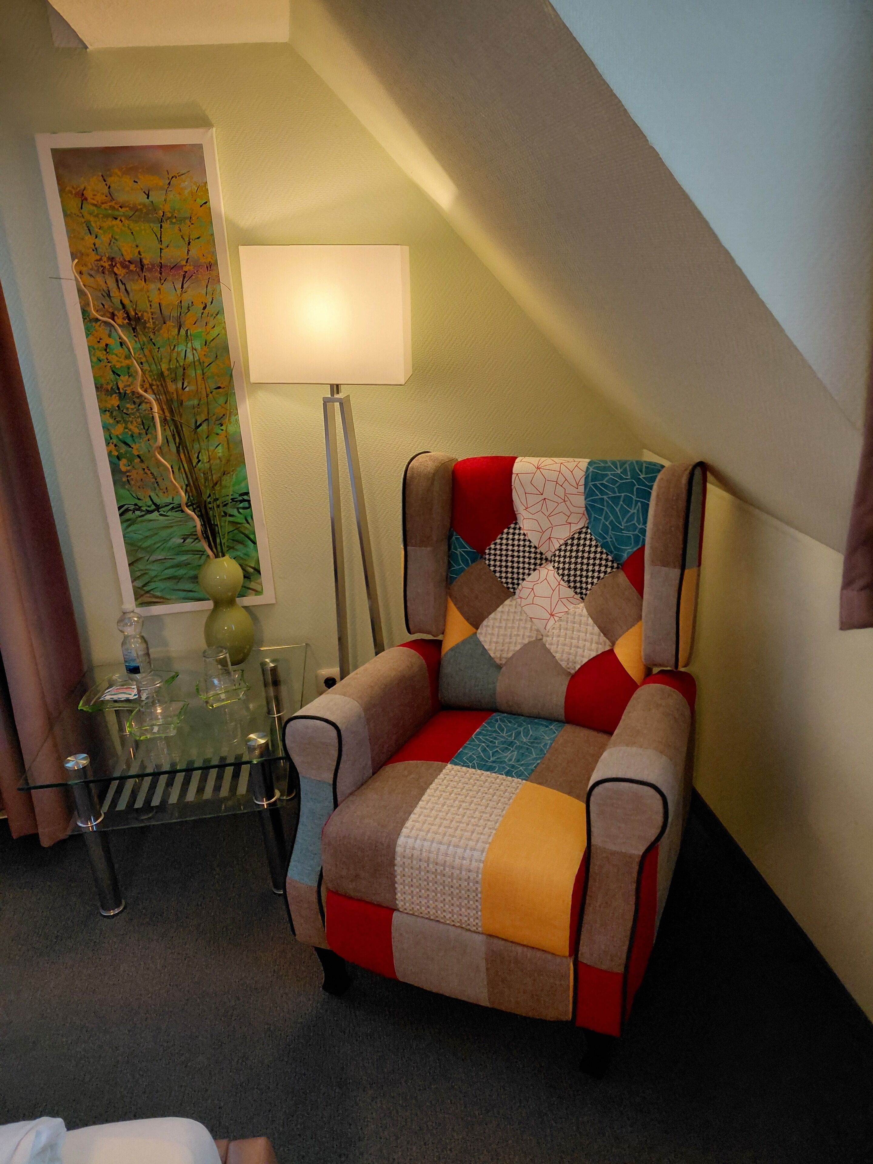 Une chambre d’hôtel chaleureuse, avec un fauteuil coloré, une table en verre ornée d’un vase et un tableau accroché au mur.