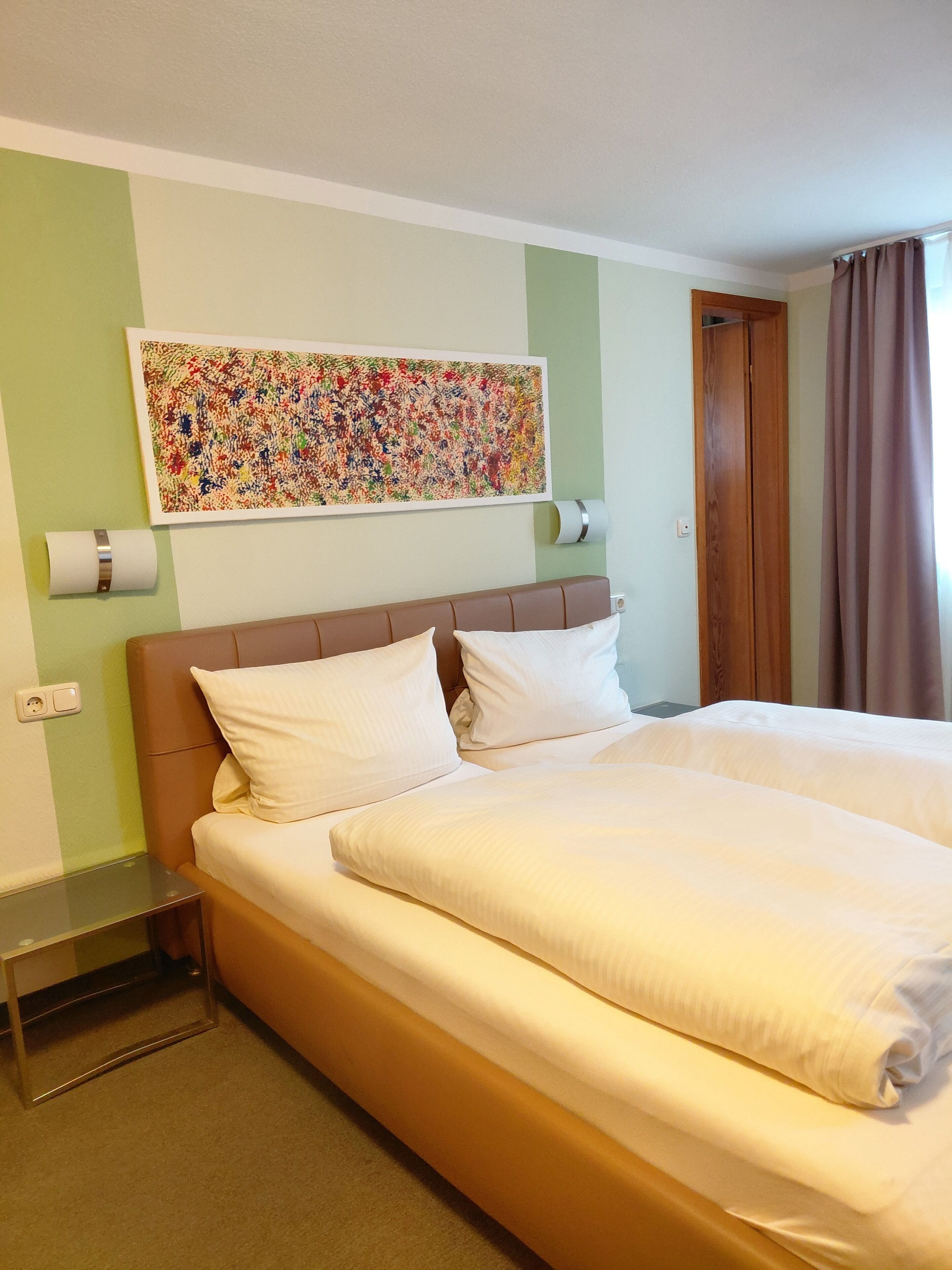 A hotel room with a double bed, a wooden headboard, a colorful abstract painting on the wall, a wooden door, and a window with curtains.