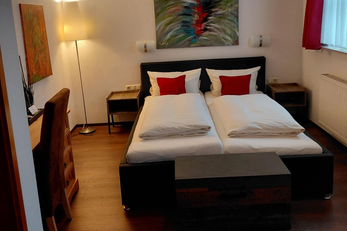 A hotel room with a large bed, two bedside tables, a desk, a chair, a floor lamp, and a painting on the wall.