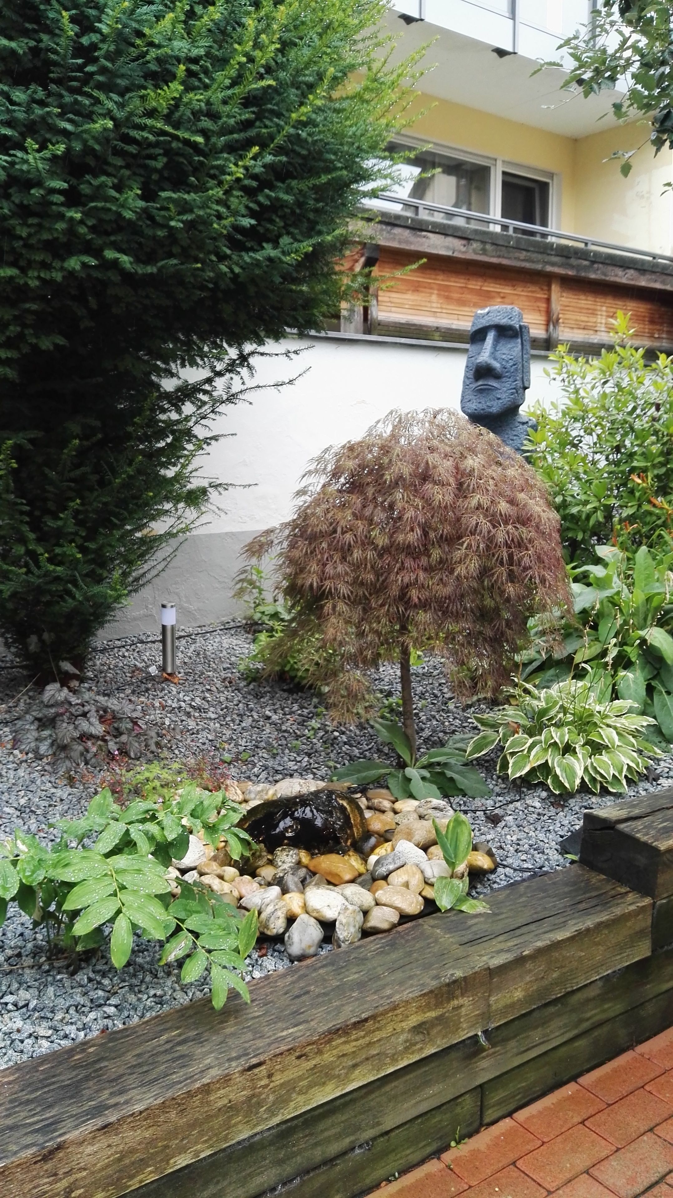 Un jardín con un pequeño estanque, una estatua y diversas plantas.