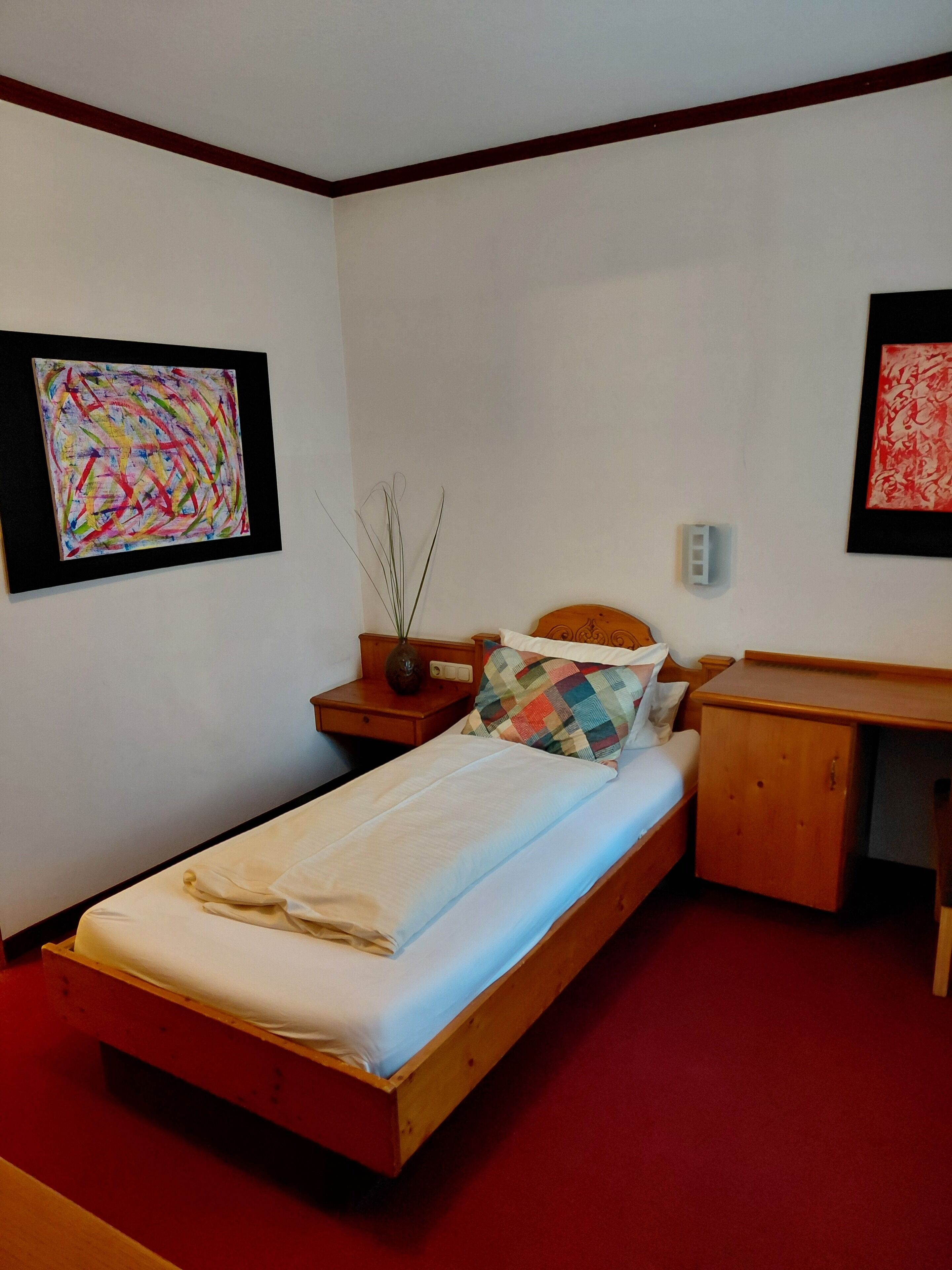Een enkel bed met houten frame, een nachtkastje met vaas, een bureau en twee abstracte schilderijen aan de muur.