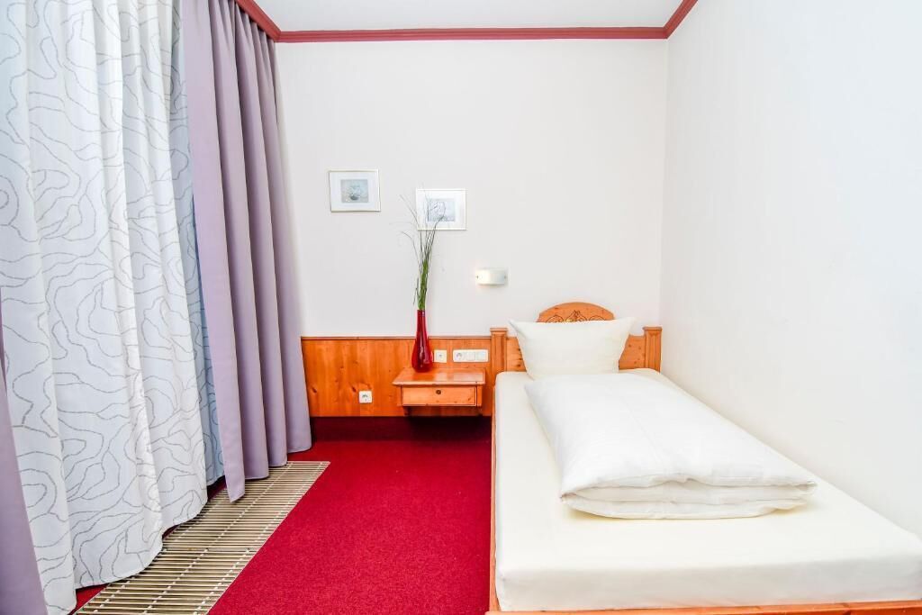 A small, neatly made room with a single bed, a wooden nightstand, a red carpet, and a wall-mounted light.