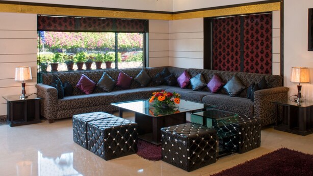 A spacious living room with a large sectional sofa, ottomans, a glass coffee table, and a chandelier. - Anila Hotel (New Delhi)