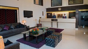 A modern living room with a sofa, coffee table, and ottomans. There is a TV mounted on the wall and a reception desk with a clock. - Anila Hotel (New Delhi)