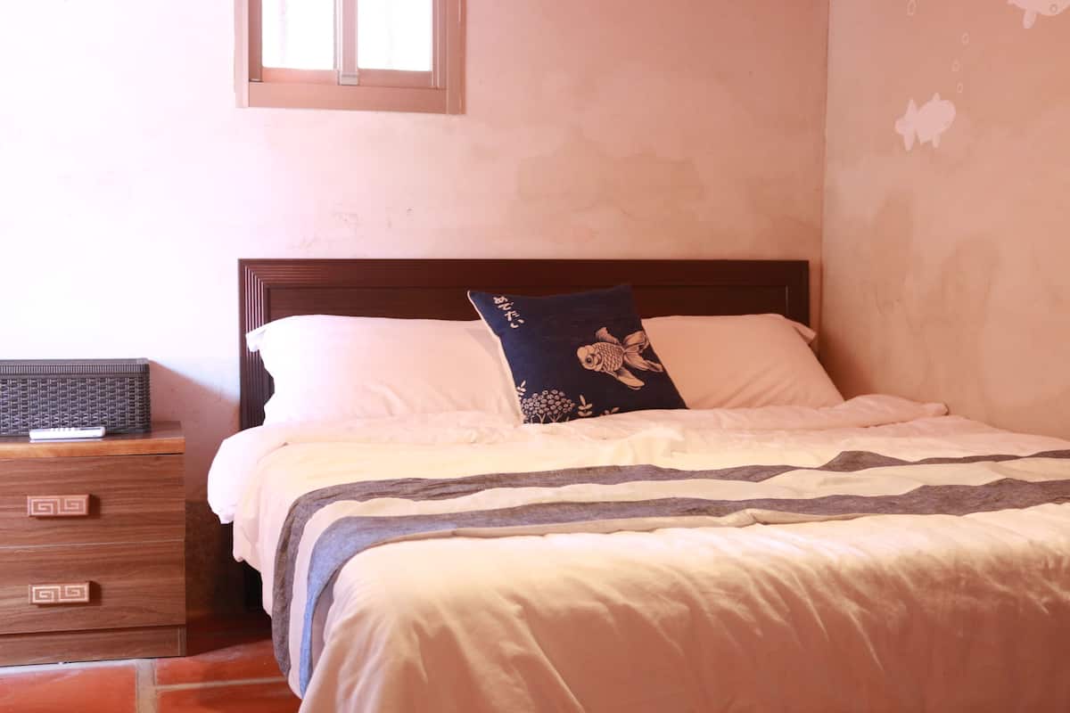 Una cama bien tendida con una colcha a rayas y un cojín decorativo, una mesita de noche de madera con cajones, y una ventana sobre la cama.