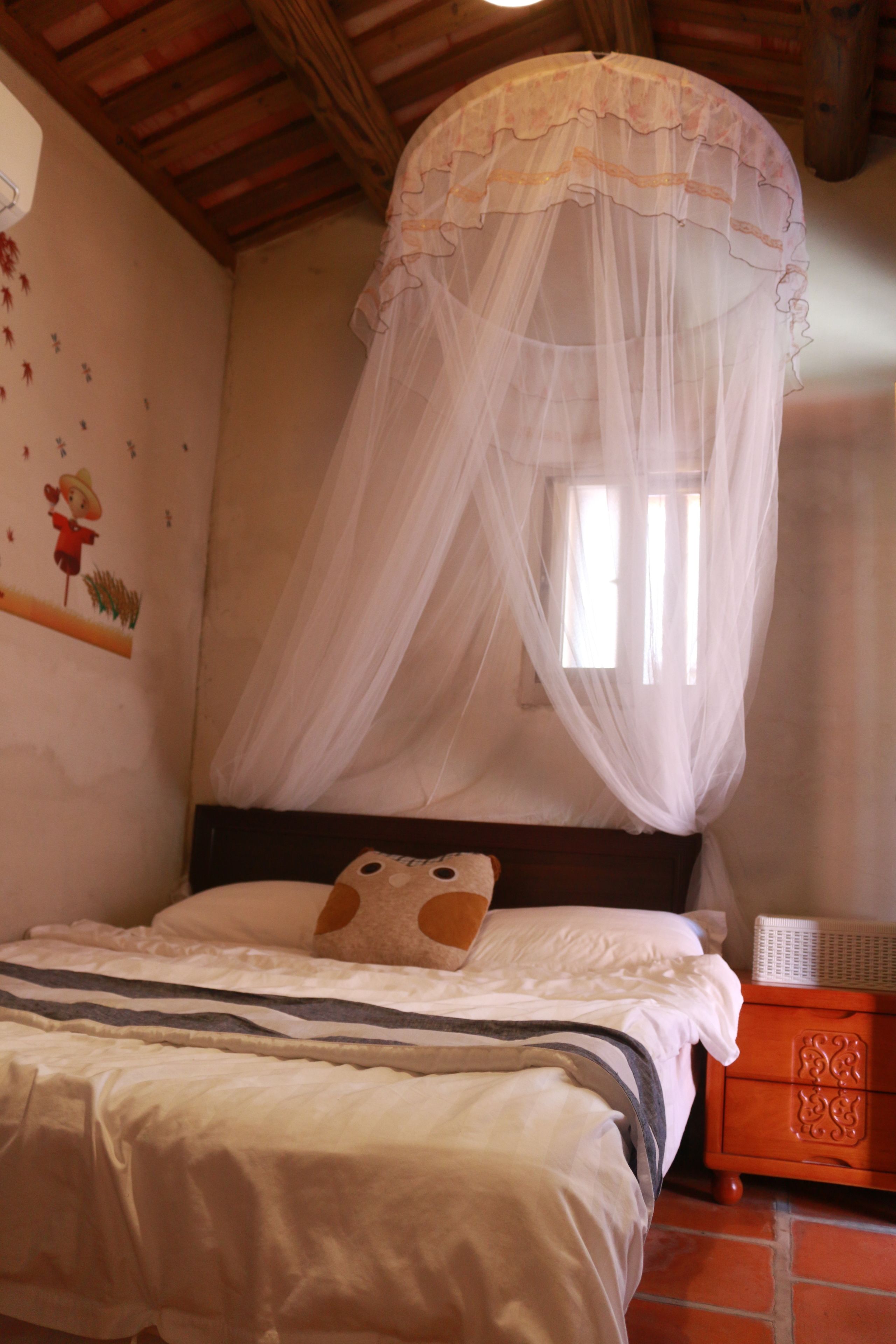 Un dormitorio con cama, mesita de noche, cabecero de madera, ventana con vista y pared con un adhesivo decorativo.