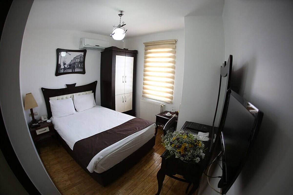 Une chambre d’hôtel comprenant un lit, un bureau avec un ordinateur, une télévision, une fenêtre avec des stores, un climatiseur et un tableau encadré au mur.