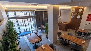 Ein Hotel-Empfangsbereich mit einem Holzbüro, mehreren Uhren, die verschiedene Städtezonen anzeigen, und einer Sitzecke mit Tischen und Stühlen.