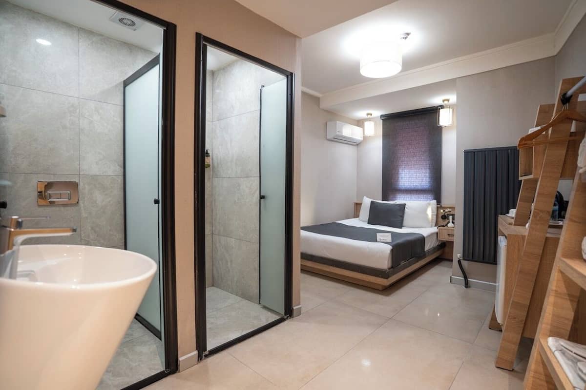 A modern hotel room with a glass-enclosed shower, a large white sink, a bed with a dark headboard, and a wooden wardrobe.