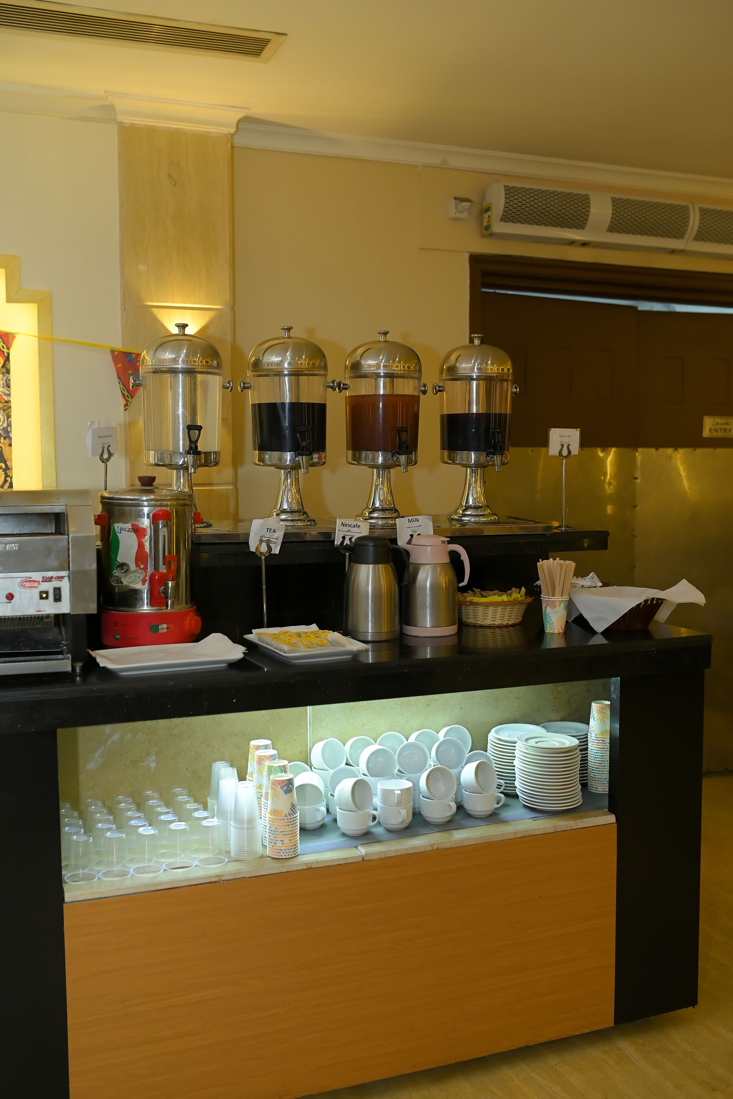 Una barra de buffet con máquinas de café, dispensadores de té y una variedad de platos y cubiertos.
