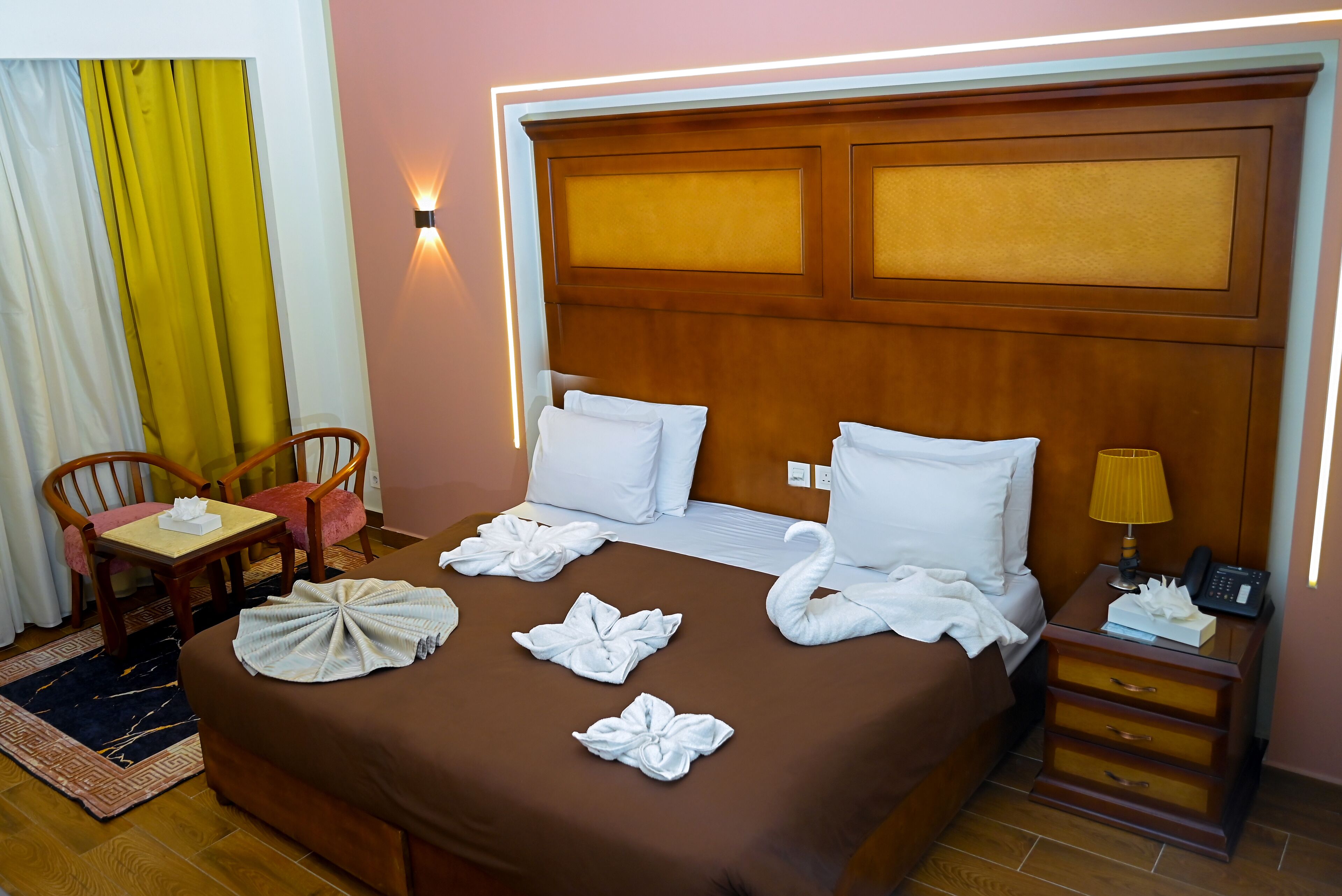 Une chambre d’hôtel avec un lit marron, des oreillers blancs, une table de chevet en bois et une petite table avec une chaise.