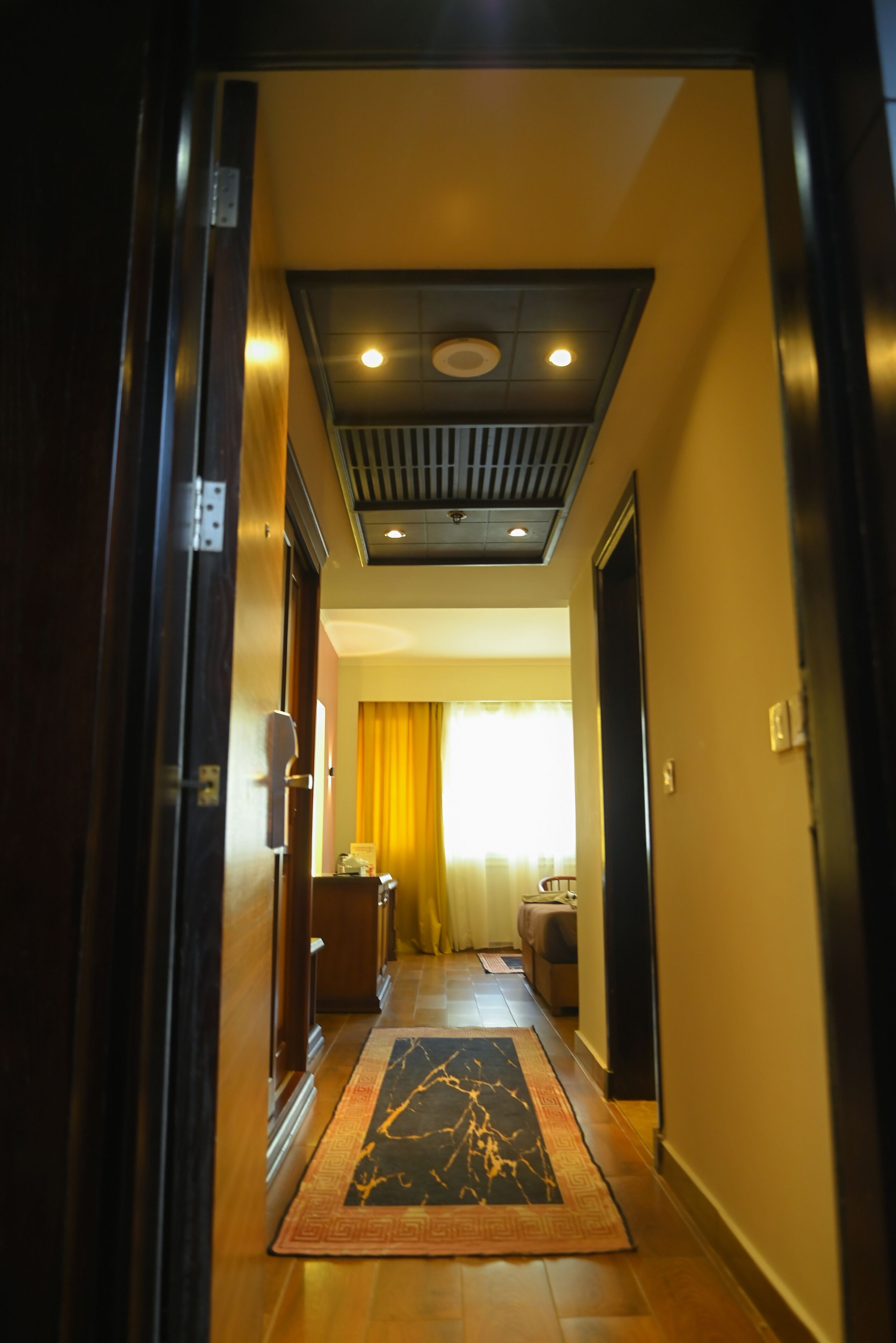 A hallway with a wooden floor, a patterned rug, a ceiling with recessed lighting, and a window with yellow curtains.