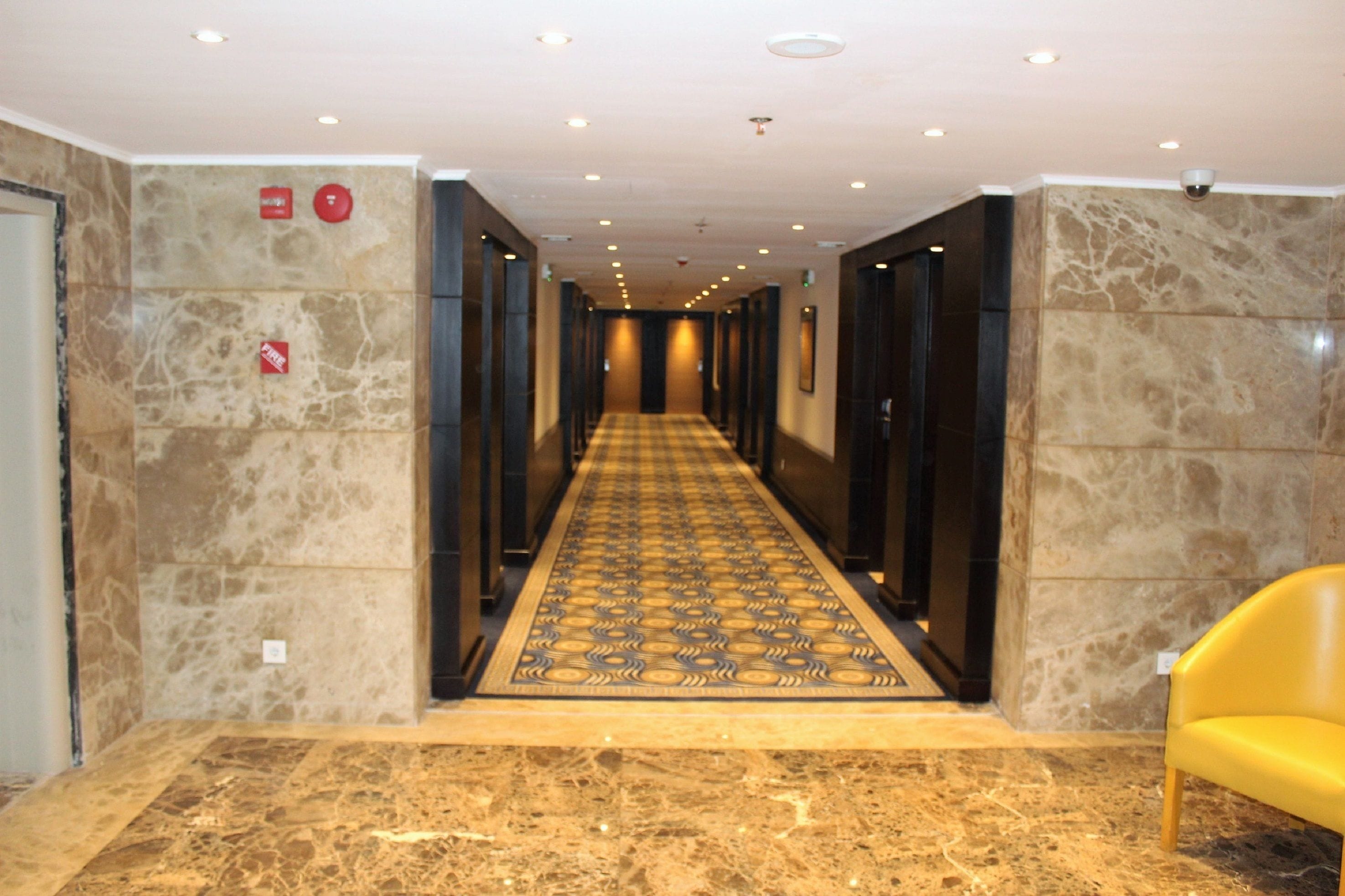 A hotel hallway with a checkered carpet, marble walls, and a yellow chair.