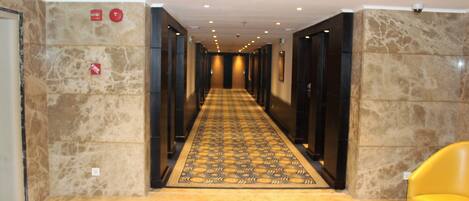 A hotel hallway with a checkered carpet, marble walls, and a yellow chair.