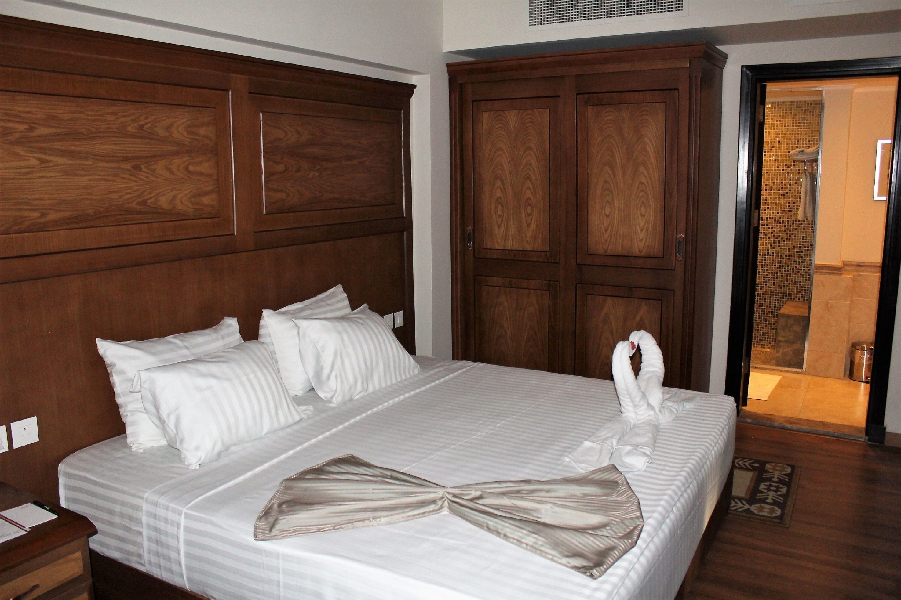Ein Hotelzimmer mit einem großen, hölzernen Kopfteil, einem ordentlich bezogenen Bett mit weißen Leinen und einer auf dem Bett drapierten Handtuchgarnitur.